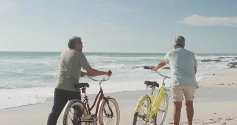 Back view of hispanic senior couple walking with bikes on beach at sunset Stock Footage 168349591