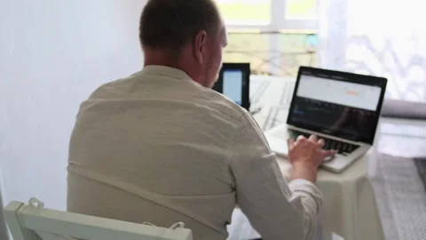 Back view of light-colored shirt man who works at home on two laptops. Stock Footage 245184430