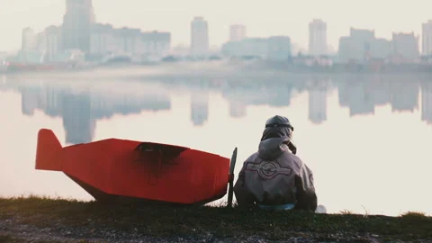 Back view of little boy in old pilot costume sitting near amazing city lake with Stock Footage 99693386