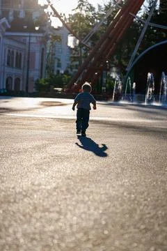 Back view of little boy running in sunny amusement park near fountains, happy ch Stock Photos