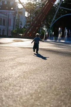 Back view of little boy running in sunny amusement park near fountains, happy ch Stock Photos