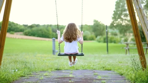 Back view of a little long-haired girl, swinging on a swing in a green meadow Stock Footage 133996563