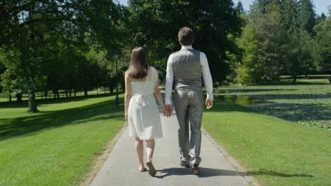 BACK VIEW: Loving couple strolling through the lovely green park hand in hand Stock Photos