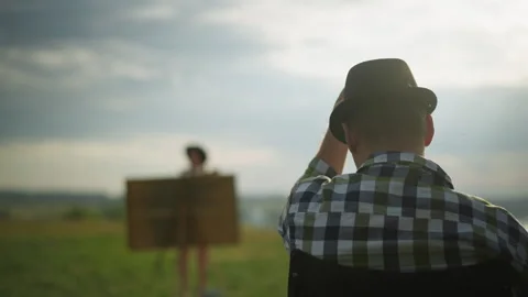 Back View of Man in Hat Seated in Field with Woman Painting in Background Stock Footage 285578490