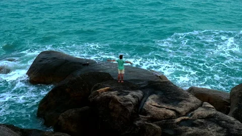 Back view of a man with raised hands standing on a stone and watching the ocean Stock Footage 132315401