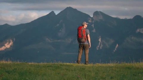 Back view man standing on hill top overlooking epic mountain peaks cloudy sky Stock Footage 283500339