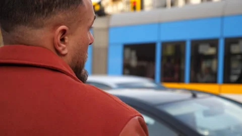 Back view of man in terracotta red jacket looking at blue city tram Stock Footage 325813014