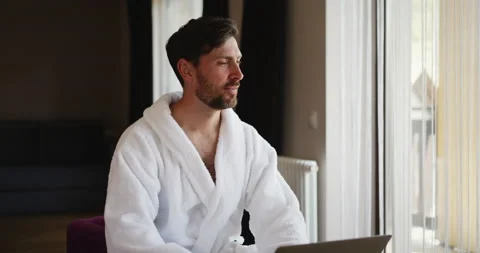 Back view of a man in white bathrobe going to the window to open the curtains in Video stock 156968593