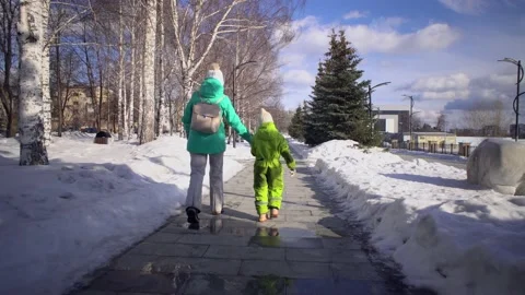 Back view of mom and kid walk hand in hand on sidewalk with snowdrifts in winter Stock Footage 140580488