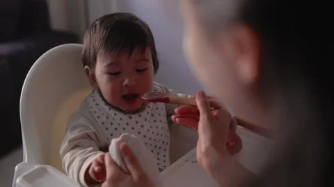 Back view of mom feeding her baby. Toddler is sitting on a high chair and eating Stock Footage 244827940