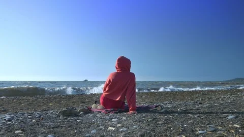 Back view. someone in a red sweatshirt and hood is sitting on the beach during a Video stock 118404270