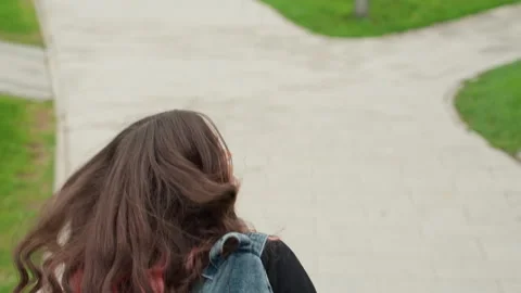 Back view student walking campus path, quiet wet sidewalk, denim backpack strap Video stock 329886634