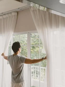 Back view of Thai man opening curtains with both hands to welcome daylight Stock-Fotos
