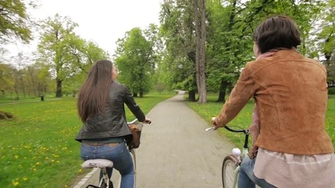 Back View of Two Brunettes in Jackets and Jeans Riding on Bicycles in the Park Stock Footage 76252317