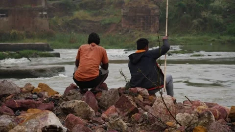 Back view of Two men sitting on rocks by the river holding Video stock 225741412
