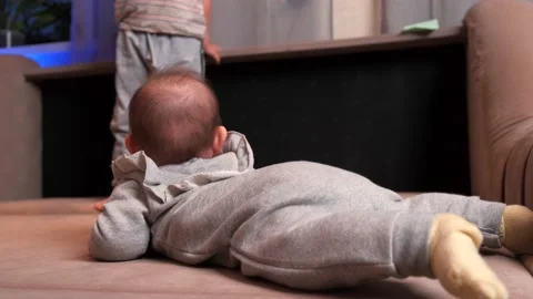 Back view video of a small newborn baby lying on a couch looking at his bigger Stock Footage 155372833