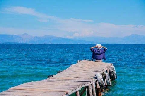 Back view of a woman sitting on a tiny wooden pier by the sea. Young beautiful Stock Photos