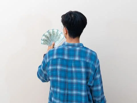 Back view of a young Asian man holding a fan of US dollar bills against a p.. Stock Photos