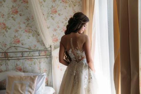 Back view of young bride in beautiful white wedding dress stands in her room in Stock Photos
