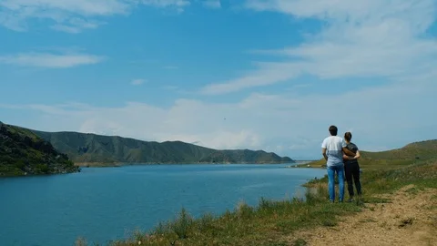 Back view of young couple standing on the shore of lake. Man hugs woman gently Видео 94425946