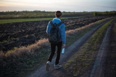 Back view of young guy with backpack walking on village road with eco bottle. Stock Photos
