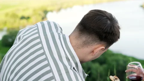 Back view. Young guy in a striped shirt eats during a picnic in nature. Vídeos de archivo 162194374