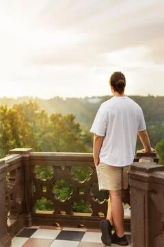 Back view of a young man standing on a stone balcony, looking out at a scen.. Stock Photos