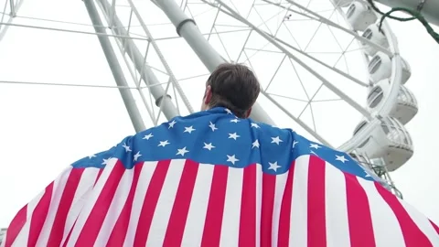 Back view Young man waving national USA flag outdoors over ferris wheel at Stock Footage 154972629
