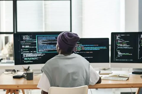 Back view of young software developer sitting in front of three monitors Stock Photos
