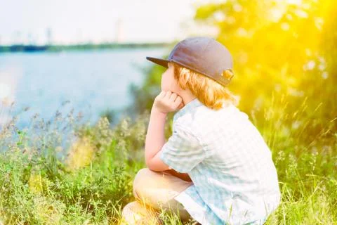 Back of young boy siting on grass facing to lake view, watching sunset in park Stock Photos