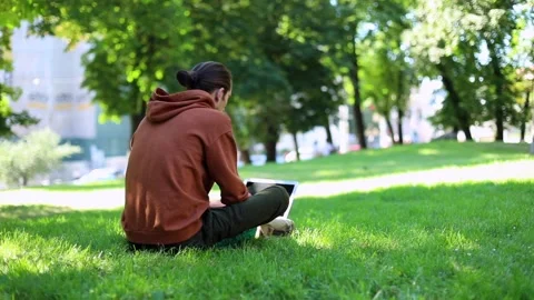 Back of young male person using modern laptop outdoor in sunny green park 스톡 동영상 139201231