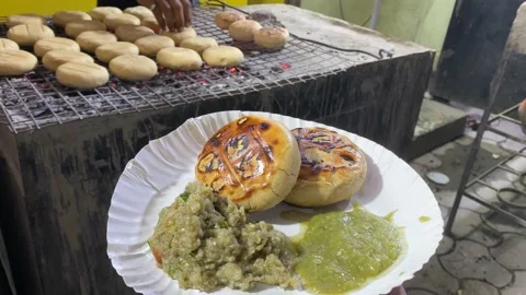 In the backdrop, a vendor is seen baking the Litti in a hot iron grill Stock Footage 205586516