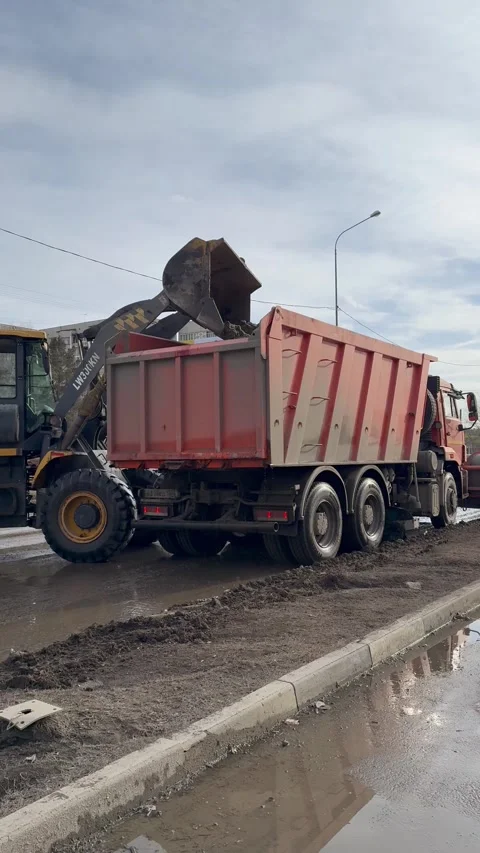 Backhoe loader unloads icy mud from city road into dump truck for removal Stock Footage 306029539