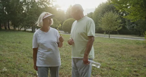Backlit elderly couple walking in a park chatting and carrying a bottle of water Video stock 134207137