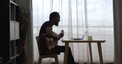 Backlit middle-aged man at home learning guitar using his laptop seated at a Stock Footage 155276749