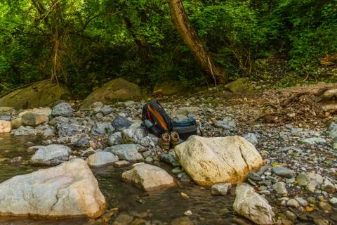 A backpack, camera bag and hiking boots on a riverbank of a mountain river du Stock Photos