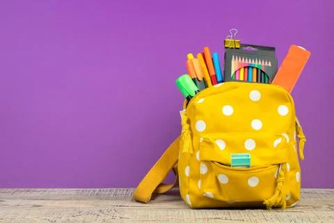 Backpack with different colorful stationery on table. Purple background. Back Stock Photos