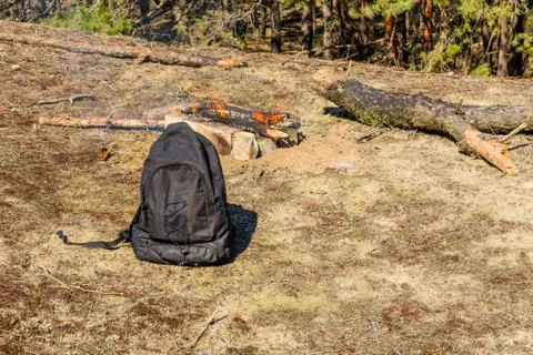 Backpack on a ground in a coniferous forest. Bonfire on a background Stock Photos