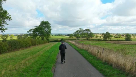 Backpacker walking along rural road between green fields and hedgerows. Active Stock Footage 346605052
