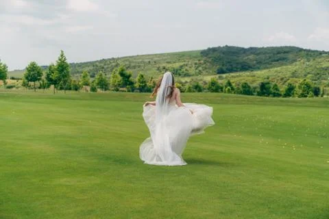 Backside view, bride runs in white wedding dress with veil on green field Stock Photos