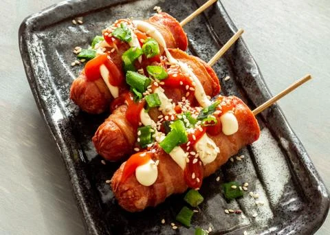 Bacon wrapped hot dogs on a stick, served with mayo and ketchup. Stock Photos