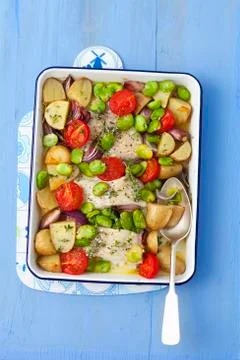 Baked cod with potatoes, broad beans, garlic, onions and tomatoes Stock Photos
