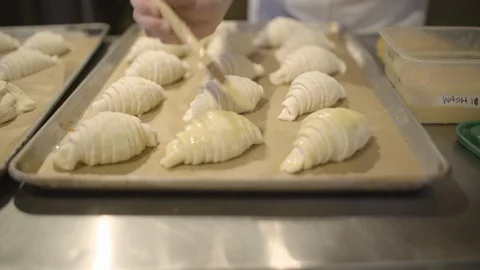 Baker applying egg wash over the rolled croissant dough with a brush Stock Footage 87612999