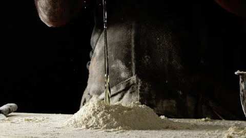 Baker in black apron cracks egg onto flour heap on counter surface black ba.. Stock Footage 274689213