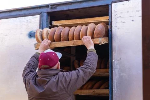 Baker in a cap unloading fresh round loaves of artisan bread from a metal d.. Stock Photos