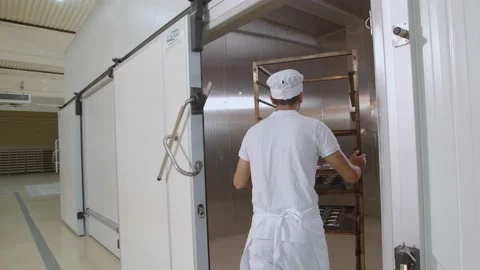 Baker dressed in white and toque puts a cart with bread into big oven Stock Footage 153467038