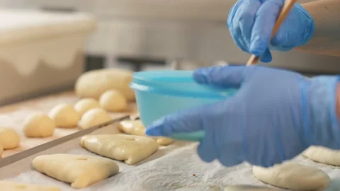 Baker hand inflicts egg mixture with brush on patties before baking on table Stock Footage 90680874