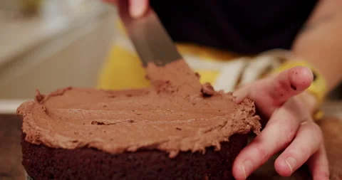 Baker icing the top of a chocolate cake with chocolate frosting in yellow apron Stock Footage 247785787