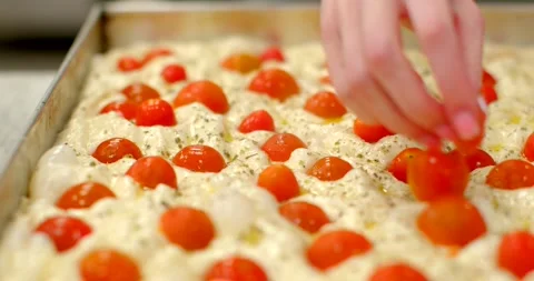 Baker placing cherry tomato halves into dimpled focaccia dough in metal tray Stock Footage 344582727