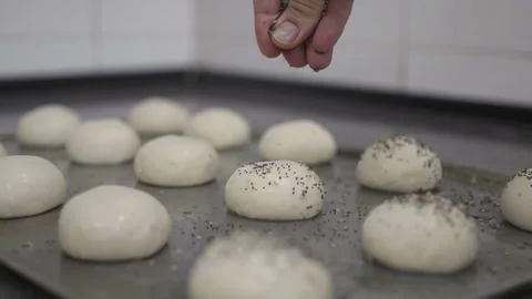 Baker sprinkling a tray of bread rolls with seeds with dough in the background 스톡 동영상 251363667